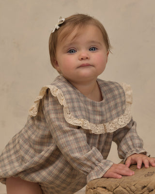 Noralee Noelle Romper - Fall Plaid
A toddler girl wearing a beige and brown plaid long-sleeve romper with a large cream lace ruffle collar, posing on a textured stone surface against a neutral beige background.