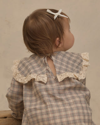 Noralee Noelle Romper - Fall Plaid
A toddler seen from behind wearing a beige and brown plaid long-sleeve top with cream lace ruffles and a white bow in their hair, set against a neutral beige background.