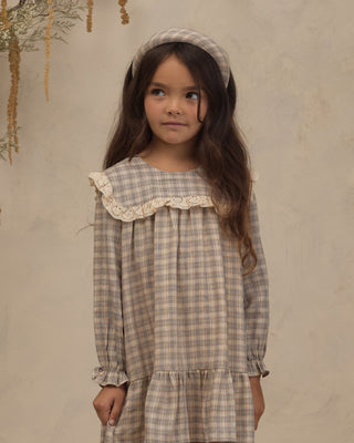 Noralee Noelle Dress - Fall Plaid
A young girl with long brown hair wearing a beige and gray plaid dress with a large cream lace ruffle collar, a matching plaid headband, and a pleated skirt, standing against a textured beige wall.