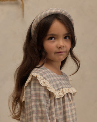 Noralee Noelle Dress - Fall Plaid
A young girl with long dark brown hair wearing a beige and brown plaid headband and a matching plaid top with a cream lace ruffle collar, photographed against a neutral textured beige wall.