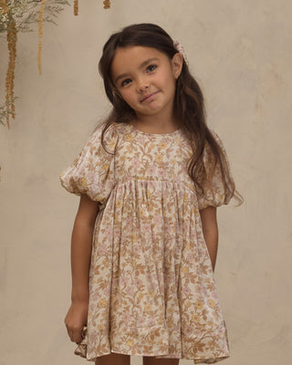 Noralee Luna Dress - Wildflower
A young girl with long dark brown hair wearing a short-sleeved, puff-sleeve dress with a beige, pink, and gold floral pattern, standing against a textured beige wall.