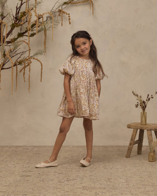 Noralee Luna Dress - Wildflower
A young girl with long dark hair stands in a studio setting wearing a short-sleeved, flutter-sleeve babydoll dress with a beige and white floral pattern. She is wearing light-colored ballet flats and posing in front of a textured beige wall decorated with dried botanicals.