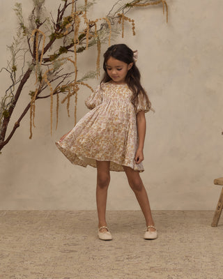 Noralee Luna Dress - Wildflower
A young girl with long dark hair stands in a studio wearing a short cream dress with a yellow floral print and flutter sleeves. She is posed in front of a textured beige wall with decorative dried branches on the left.