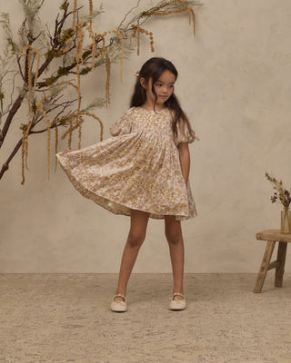 Noralee Luna Dress - Wildflower
A young girl with long dark hair stands in a studio setting wearing a short, flutter-sleeve dress with a beige and yellow floral pattern. She is twirling, causing the skirt of the dress to flare out.