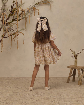 Noralee Alice Bow - Wildflower
Noralee Luna Dress - Wildflower
A young girl is shown from behind wearing a short cream-colored floral dress with a matching fabric bow headband. She is standing in a rustic room with a textured beige wall, a small wooden stool, and dried botanical decorations.