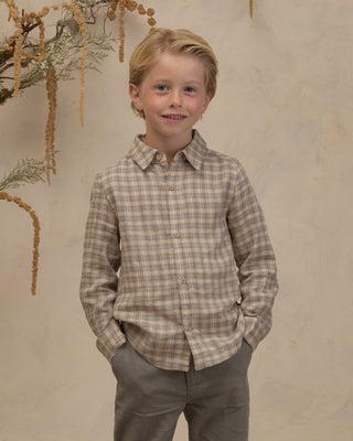 Noralee Harrison Button Down - Fall Plaid
A young boy with blonde hair stands wearing a long-sleeve button-up plaid shirt in beige and brown tones, paired with gray trousers, against a textured beige wall with dried plant decorations.