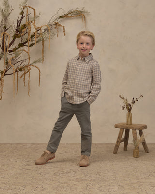Noralee Harrison Button Down - Fall Plaid
A young boy stands in a studio setting wearing a beige and brown plaid button-down shirt, gray trousers, and tan shoes. The background features a textured beige wall with dried botanical decorations and a small wooden stool.