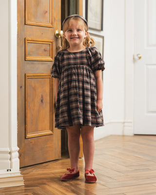 Noralee Luna Dress - Tartan
A young girl stands in a doorway wearing a short-sleeve plaid dress with puff sleeves, paired with red shoes and a dark headband. The dress features a brown, black, and beige plaid pattern.
