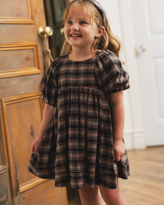 Noralee Luna Dress - Tartan
A young girl with blonde hair wearing a brown and black plaid dress, standing in front of a wooden door and white wall.