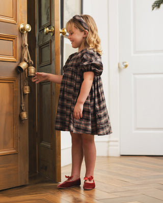Noralee Luna Dress - Tartan
A young girl with blonde hair stands in a doorway, ringing a brass door bell. She is wearing a dark brown and beige plaid puff-sleeve dress with a matching headband and dark red moccasin-style shoes.