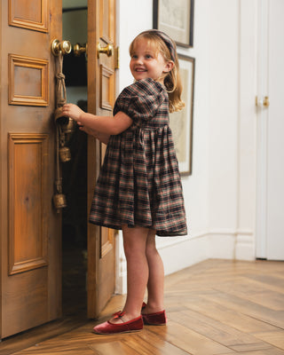 Noralee Luna Dress - Tartan
A young girl with blonde hair stands in a doorway wearing a brown and black plaid dress and red shoes, holding the door open with one hand.