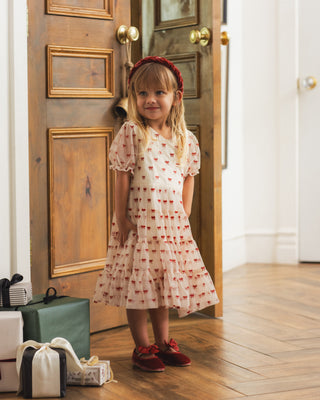 Noralee Dottie Dress - Ruby Bows
A young girl with blonde hair wearing a red bow headband, a cream puff-sleeve dress with a red heart pattern, and red shoes, standing in front of an open wooden door in a home interior.