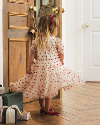 Noralee Dottie Dress - Ruby Bows
A young girl with blonde hair and a red headband is seen from behind, wearing a light pink flutter-sleeve dress with a red floral pattern and red shoes, standing in a room with a wooden door and herringbone wood floor.
