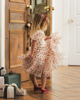Noralee Dottie Dress - Ruby Bows
A young girl with blonde hair wearing a white tulle dress with red floral patterns and red shoes, standing in a doorway and holding out the skirt of the dress. She is in a home interior with wooden floors and a wooden door.