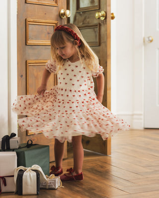 Noralee Dottie Dress - Ruby Bows
A young girl with blonde hair and a red headband twirls in a white tulle dress with red embroidered stars, standing in a doorway with wooden floors and gift boxes at her side.