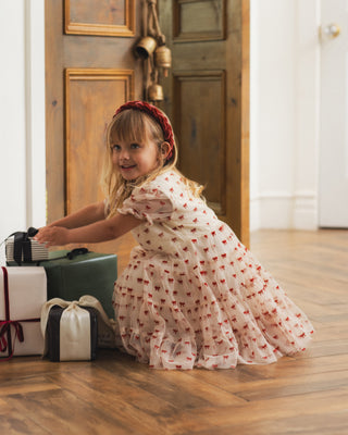 Noralee Dottie Dress - Ruby Bows
A young girl with a red headband kneels on a wooden floor next to wrapped gifts, wearing a white tulle dress with a red cherry print.