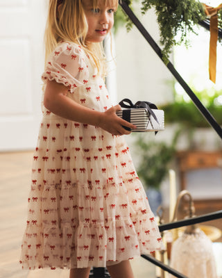 Noralee Dottie Dress - Ruby Bows
A young girl with blonde hair wearing a tiered cream tulle dress with a red bow pattern, holding a striped gift box with a black ribbon, standing in a decorated indoor setting.