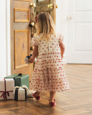 Noralee Dottie Dress - Ruby Bows
A young girl with blonde hair is seen from behind wearing a cream-colored short-sleeve dress with a red pattern, walking on a wooden floor in a home interior next to wrapped gifts.