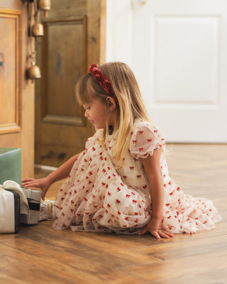 Noralee Dottie Dress - Ruby Bows
A young girl with blonde hair sits on a wooden floor wearing a white tulle dress with red bow patterns and a red bow headband, looking down at a small striped box.