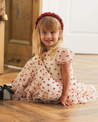 Noralee Dottie Dress - Ruby Bows
A young girl with blonde hair sits on a wooden floor wearing a white tulle dress with a red bow pattern and a red headband. A small striped gift box is on the floor next to her.