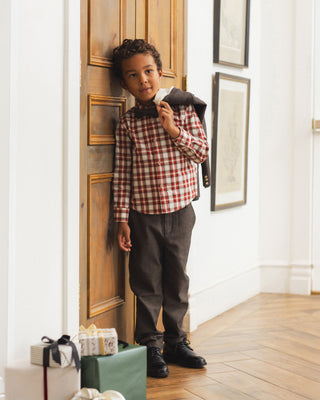 Noralee Harrison Button Down - Holiday Tartan
A young boy stands in a hallway leaning against a wooden door, wearing a red and white gingham long-sleeve shirt, a dark bow tie, dark trousers, and black shoes, while holding a dark jacket over his shoulder.