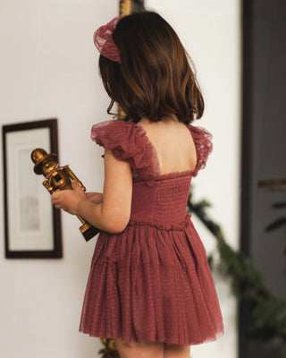 Noralee Gigi Tutu - Ruby 
A young girl viewed from behind wearing a dusty rose puff-sleeve tulle dress and matching hair accessory, holding a vintage-style brass toy in an indoor setting.