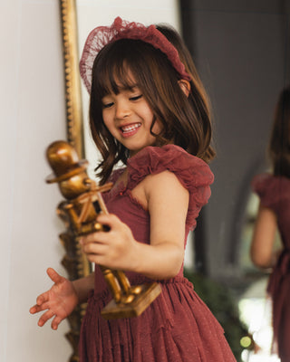 Noralee Gigi Tutu - Ruby
A young girl with brown hair wearing a dusty rose ruffled headband and matching ruffled dress, smiling while holding a golden nutcracker toy in an indoor setting.