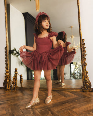 Noralee Gigi Tutu - Ruby
A young girl wearing a dusty rose ballet dress with a matching bonnet and metallic ballet shoes, posing in front of a large ornate gold mirror that reflects her pose.
