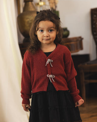 Noralee Bow Cardigan - Ruby
A young girl with curly brown hair wearing a red knit cardigan with white cord ties and a black dress, standing in a softly lit indoor room.