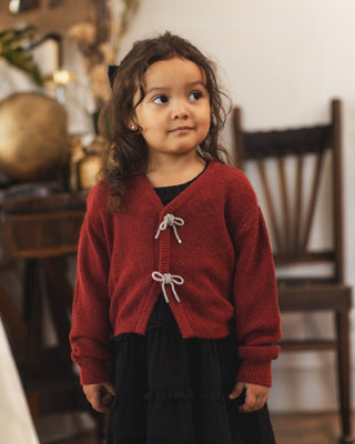 Noralee Bow Cardigan - Ruby
A young girl with long brown hair wearing a red knit cardigan with white cord ties over a black outfit, standing in a softly blurred indoor setting with a wooden chair in the background.