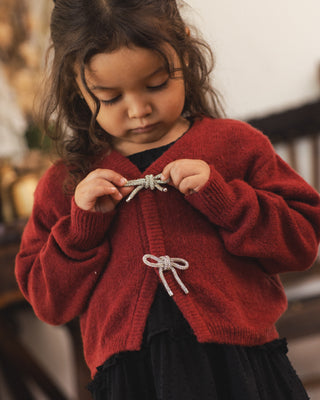 Noralee Bow Cardigan - Ruby
A young child wearing a red knit cardigan with silver bow details, standing in a softly blurred indoor setting.