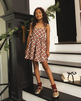 Noralee Betsy Dress - Holiday Tartan
A young girl stands on an indoor staircase wearing a red and white gingham mini dress with a tie waist, red socks, and metallic shoes. The background features a dark wooden banister, white walls, and holiday greenery.