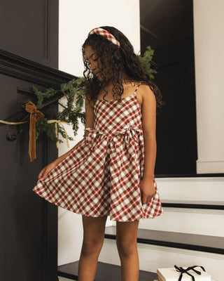 Noralee Betsy Dress - Holiday Tartan
A young girl with curly dark hair wearing a red and white gingham check dress and matching headband, standing on white stairs in a home interior next to a dark door with holiday greenery.