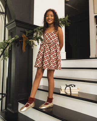 Noralee Betsy Dress - Holiday Tartan
A young girl stands on indoor stairs wearing a red and white gingham mini dress with thin straps and a gathered waist. She is styled with red socks and metallic gold flats, with holiday greenery and a dark paneled interior in the background.