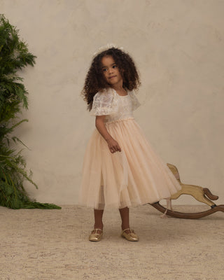 Noralee Dorothy Dress - Pearl
A young girl with curly dark hair stands indoors wearing a formal dress with a white lace bodice and a soft blush-beige tulle skirt. She is posed in front of a textured beige wall, with a green plant on the left and a wooden rocking horse on the right.