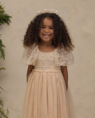 Noralee Dorothy Dress - Pearl
A young girl with curly brown hair smiling while wearing a light peach tulle dress with sheer puff sleeves and a matching headpiece, set against a textured beige wall with green foliage on the side.