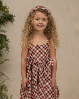 Noralee Padded Headband - Holiday Tartan
Noralee Betsy Dress - Holiday Tartan
A young girl with curly blonde hair wearing a red and beige gingham dress with a matching headband, standing against a textured beige wall with green foliage on the left.