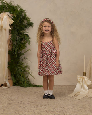 Noralee Betsy Dress - Holiday Tartan
A young girl with curly blonde hair stands in a studio setting wearing a red and white gingham strapless dress with side bows, a matching gingham headband, white socks, and black Mary Jane shoes. The background features a textured beige wall, a green pine garland with a cream ribbon on the left, and a cream satin bow and candle holder on the right.