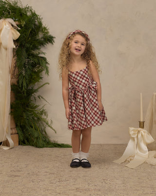 Noralee Betsy Dress - Holiday Tartan
A young girl with curly blonde hair stands in a studio wearing a red and white gingham check strapless dress with a matching headband, white socks, and black Mary Jane shoes. The background features a textured beige wall, a green foliage arrangement on the left, and a cream ribbon and candle prop on the right.
