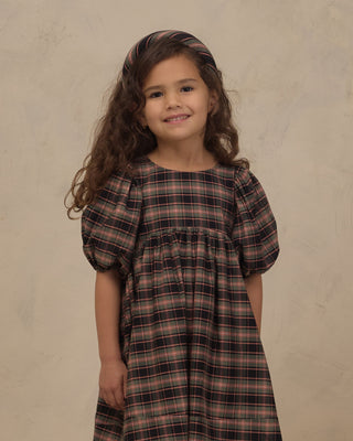 Noralee Luna Dress - Tartan
A young girl with long, wavy brown hair wearing a dark brown and black plaid dress with puff sleeves and a matching headband, standing against a textured beige background.