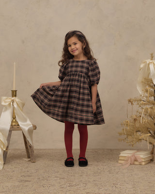 Noralee Luna Dress - Tartan
A young girl with curly brown hair stands in a studio setting wearing a short puff-sleeve plaid dress in dark brown and black, paired with dark red tights and black shoes. She is posing with a dress form on the left and a decorative dried floral arrangement with books on the right, against a textured beige backdrop.