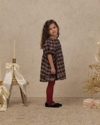 Noralee Luna Dress - Tartan
A young girl stands in a studio setting wearing a dark brown and black plaid dress with red tights and black shoes. The background features a textured beige wall and floor, decorated with a small gold holiday tree on the right and a white satin bow on a wooden stand on the left.