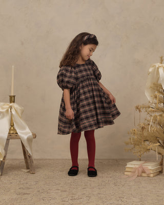Noralee Luna Dress - Tartan
A young girl stands in a studio setting wearing a dark brown and black plaid dress with puff sleeves, red tights, and black shoes. The background features a textured beige wall, a small decorated Christmas tree with gold ornaments, and a rustic wooden stool with a white ribbon and candle.