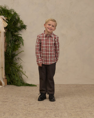 Noralee Harrison Button Down - Holiday Tartan
A young boy stands in a studio wearing a red and white plaid button-down shirt, dark brown pants, and black shoes against a textured beige backdrop with greenery on the left.