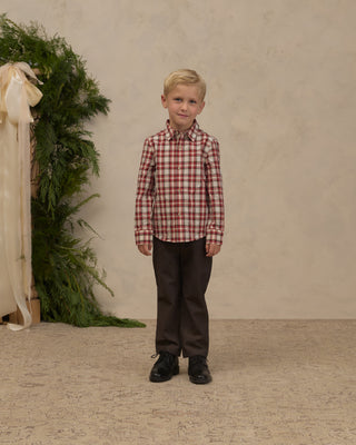 Noralee Harrison Button Down - Holiday Tartan
A young boy stands facing forward in a studio setting, wearing a red and white gingham long-sleeve button-down shirt, dark brown trousers, and black shoes. The background is a textured beige wall with a green foliage arrangement on the left.
