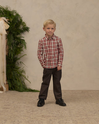 Noralee Harrison Button Down - Holiday Tartan
A young blond boy stands in a studio wearing a red and white plaid long-sleeve button-down shirt, dark brown trousers, and black shoes. The background features a textured beige wall, a patterned beige carpet, and a decorative greenery arrangement on the left.