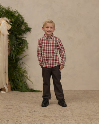 Noralee Harrison Button Down - Holiday Tartan 
A young boy stands in a studio wearing a red and white checkered long-sleeve button-down shirt, dark brown trousers, and black shoes. The background features a textured beige wall, a patterned beige floor, and a decorative greenery and cream fabric prop on the left.
