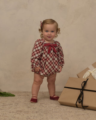Noralee Annette Romper -  Holiday Tartan
A toddler girl stands in a studio setting wearing a red and white plaid long-sleeve romper with a red bow at the neckline. She is posed next to wrapped gift boxes against a textured beige backdrop.