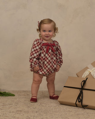 Noralee Annette Romper -  Holiday Tartan 
A toddler girl stands in a studio wearing a red and white gingham long-sleeve romper with a red bow at the neckline. She is posed next to wrapped gift boxes on a textured beige floor against a neutral beige backdrop.
