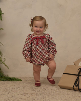 Noralee Annette Romper -  Holiday Tartan
A toddler girl wearing a red and cream plaid long-sleeve romper with a red bow at the neckline, standing on a textured beige floor against a neutral beige wall. A brown gift box is visible on the right side of the frame.